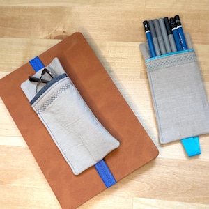 May include: A tan leather notebook with a beige fabric sleeve featuring a blue elastic band and a pair of glasses inside. Beside it, a separate beige fabric pencil case with a blue elastic band contains six pencils with blue and black accents.