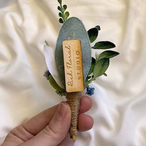 K&ouml;nnte beinhalten: Ein kleiner Strau&szlig; wei&szlig;er und blauer Blumen mit Gr&uuml;n, gebunden mit Schnur und einem Holzschild mit der Aufschrift "Rish Floral STUDIO".