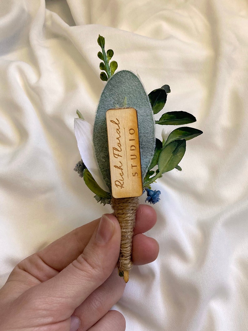 K&ouml;nnte beinhalten: Ein kleiner Strau&szlig; wei&szlig;er und blauer Blumen mit Gr&uuml;n, gebunden mit Schnur und einem Holzschild mit der Aufschrift "Rish Floral STUDIO".