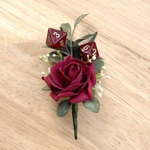 May include: A boutonniere featuring a deep red rose, green foliage, and small white floral accents. Two translucent dark red polyhedral dice with gold-colored numbers are nestled among the leaves, with one showing a three and the other a six.