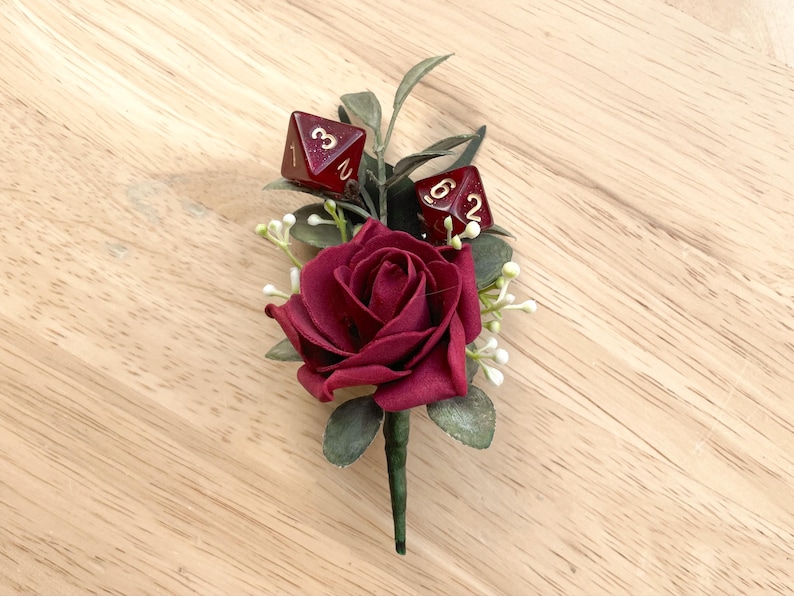 May include: A boutonniere featuring a deep red rose, green foliage, and small white floral accents. Two translucent dark red polyhedral dice with gold-colored numbers are nestled among the leaves, with one showing a three and the other a six.