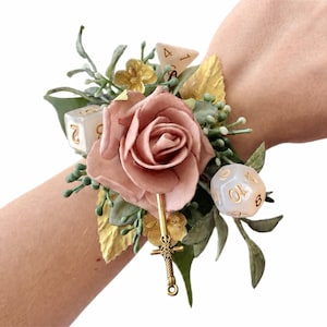 May include: A wrist corsage featuring a dusty pink artificial rose, green foliage, gold-toned leaves, and small white flowers. The arrangement is accented with two translucent polyhedral dice with gold numbers and a small gold-toned sword charm.