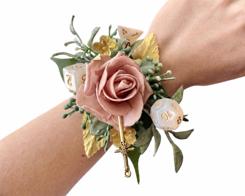 May include: A wrist corsage featuring a dusty pink artificial rose, green foliage, gold-toned leaves, and small white flowers. The arrangement is accented with two translucent polyhedral dice with gold numbers and a small gold-toned sword charm.