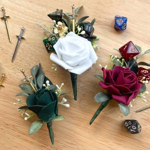 May include: Three floral boutonnieres featuring white, dark green, and burgundy roses accented with green leaves and small white berries. Each boutonniere is decorated with miniature metal sword charms and multi-sided gaming dice in various colors.