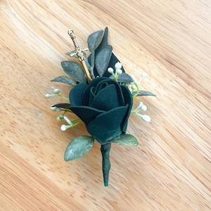 May include: A boutonniere featuring a single dark forest green rose, accented with muted green leaves and small white floral buds. A small, metallic gold-toned sword charm is tucked into the foliage, positioned vertically behind the rose bloom.
