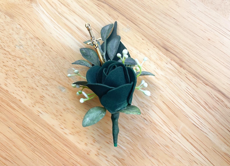 May include: A boutonniere featuring a single dark forest green rose, accented with muted green leaves and small white floral buds. A small, metallic gold-toned sword charm is tucked into the foliage, positioned vertically behind the rose bloom.