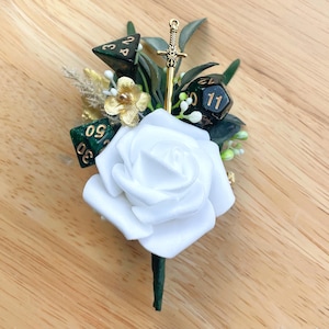 May include: A boutonniere featuring a large white foam rose, dark green foliage, and three polyhedral dice with gold numbering. A small gold-toned sword charm is positioned vertically behind the rose, accented by gold-colored floral elements.