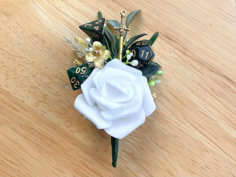 May include: A boutonniere featuring a large white foam rose, dark green foliage, and three polyhedral dice with gold numbering. A small gold-toned sword charm is positioned vertically behind the rose, accented by gold-colored floral elements.