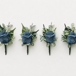 May include: Four boutonnieres featuring a dusty blue rose, green foliage, and a small silver sword charm. Each floral arrangement is wrapped in dark green floral tape, creating a cohesive set of decorative accessories for formal attire.