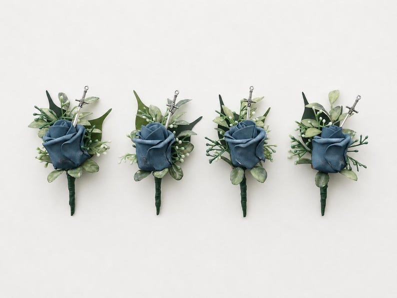 May include: Four boutonnieres featuring a dusty blue rose, green foliage, and a small silver sword charm. Each floral arrangement is wrapped in dark green floral tape, creating a cohesive set of decorative accessories for formal attire.