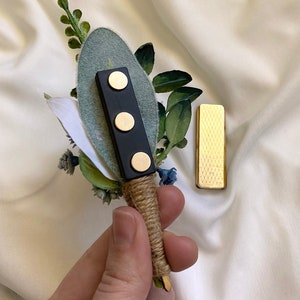 K&ouml;nnte beinhalten: Ein kleiner Strau&szlig; aus Gr&uuml;npflanzen und blauen Blumen mit einer schwarzen Magnetnadel und einer goldenen Metallnadel. Der Strau&szlig; ist mit Schnur gebunden.