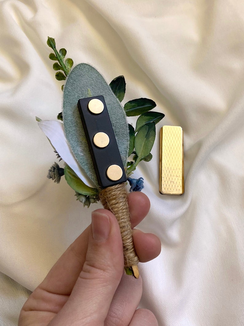 K&ouml;nnte beinhalten: Ein kleiner Strau&szlig; aus Gr&uuml;npflanzen und blauen Blumen mit einer schwarzen Magnetnadel und einer goldenen Metallnadel. Der Strau&szlig; ist mit Schnur gebunden.