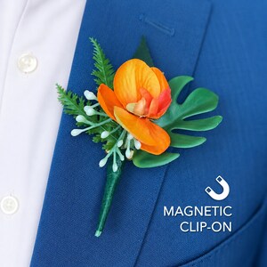 May include: A floral boutonniere featuring a bright orange orchid, a green monstera leaf, fern foliage, and small white berries. The accessory includes a magnetic clip-on mechanism, as indicated by the text MAGNETIC CLIP-ON and a magnet icon.