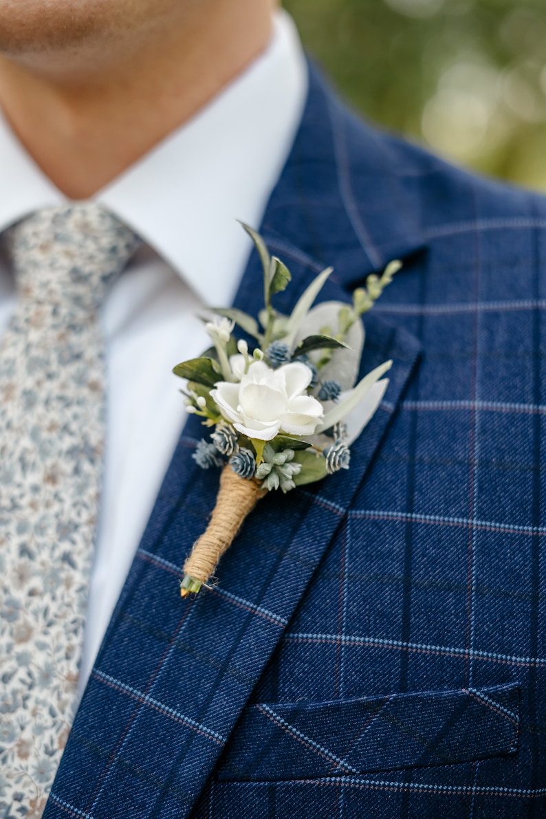 K&ouml;nnte beinhalten: Ein blaues kariertes Sakko mit einem wei&szlig;en Hemd und einem floralen Ansteckblumen.