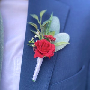 May include: A red rose boutonniere with greenery and white accents. The boutonniere is pinned to a blue suit jacket.