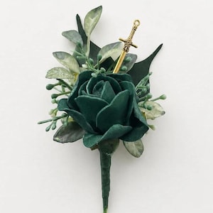 May include: A boutonniere featuring a dark green rose surrounded by light green foliage and a small gold-colored sword charm. The stem is wrapped in dark green floral tape, creating a structured floral arrangement with a metallic accent piece.