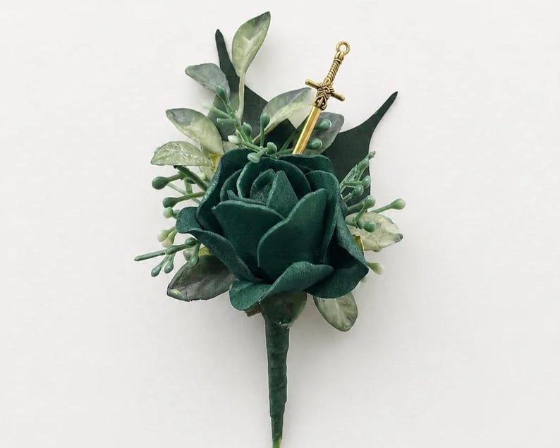 May include: A boutonniere featuring a dark green rose surrounded by light green foliage and a small gold-colored sword charm. The stem is wrapped in dark green floral tape, creating a structured floral arrangement with a metallic accent piece.