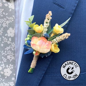 May include: A floral boutonniere with yellow, peach, and blue accents, attached to a navy blue blazer. The boutonniere is accented with greenery and wrapped with twine. A gray floral patterned tie is visible. A white and black "MAGNETIC CLIP-ON" sticker is also visible.