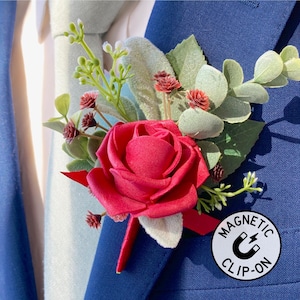 May include: A red rose boutonniere with greenery and small red accents, attached to a blue blazer. The boutonniere has a magnetic clip-on feature, as indicated by the white circular tag with the text "MAGNETIC CLIP-ON". The blazer is paired with a light blue tie and white shirt.