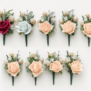 May include: A collection of nine floral boutonnieres featuring a single rose, green foliage, small white flowers, a silver sword charm, and several polyhedral dice. The roses vary in color, including deep red, light blue, and shades of peach.