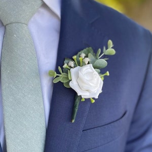 May include: A white rose boutonniere with greenery and small white flowers pinned to a navy blue suit jacket. A light green tie with a subtle pattern is visible. The image is a close-up of formal wear.
