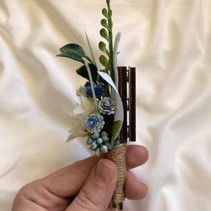 K&ouml;nnte beinhalten: Ein kleiner Strau&szlig; aus wei&szlig;en und blauen Blumen mit gr&uuml;nen Bl&auml;ttern, gebunden mit Schnur und an einer Holzbasis befestigt.