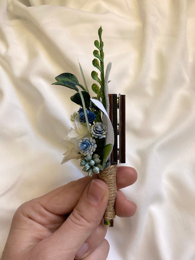 K&ouml;nnte beinhalten: Ein kleiner Strau&szlig; aus wei&szlig;en und blauen Blumen mit gr&uuml;nen Bl&auml;ttern, gebunden mit Schnur und an einer Holzbasis befestigt.