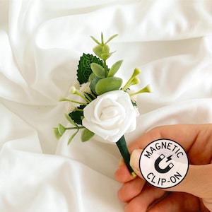 May include: White rose boutonniere with greenery and small white flowers. The boutonniere has a magnetic clip-on attachment.