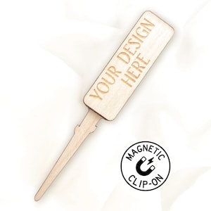May include: A light-coloured wooden sign with the text "YOUR DESIGN HERE" engraved on it. The sign has a pointed end and a decorative stem. A circular graphic with the words "MAGNETIC CLIP-ON" surrounds a magnet icon.