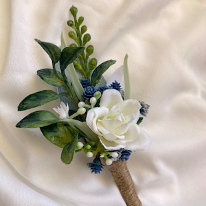 K&ouml;nnte beinhalten: Ein kleines wei&szlig;es Rosen-Boutonniere mit blauen und gr&uuml;nen Akzenten. Das Boutonniere ist mit Schnur umwickelt.