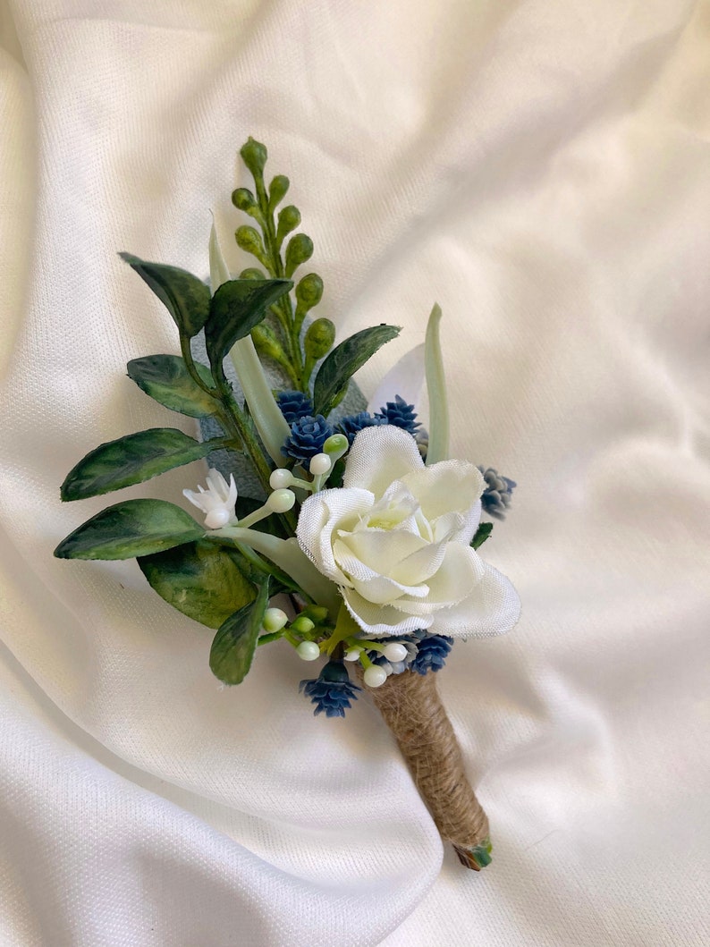 K&ouml;nnte beinhalten: Ein kleines wei&szlig;es Rosen-Boutonniere mit blauen und gr&uuml;nen Akzenten. Das Boutonniere ist mit Schnur umwickelt.
