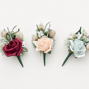 May include: Three floral boutonnieres featuring a single rose in deep red, pale peach, or light blue. Each arrangement includes green foliage, small white flowers, polyhedral dice with gold numbering, and a miniature silver sword charm.