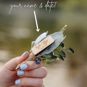 K&ouml;nnte beinhalten: Ein kleines Bouquet aus Gr&uuml;npflanzen mit einem Holzschild, auf dem "Hayden + Elyse 5-27-23" steht.