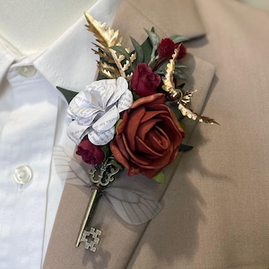 May include: A boutonniere pinned to a tan blazer and white shirt, featuring a rust-colored rose, a white paper rose made from book pages, small dark red flowers, gold-toned metallic leaves, and a vintage-style skeleton key with wings.