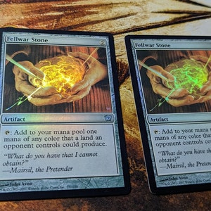 May include: Two "Fellwar Stone" trading cards. The cards have a green and yellow glowing orb graphic. The text on the cards includes the name "Fellwar Stone", the word "Artifact", and other game instructions. The cards are on a patterned surface.