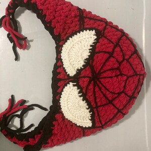May include: A handmade crochet hat in the style of Spider-Man's mask. The hat features a red base with a black web design and white eye details. Black yarn is used for the edges and ties.