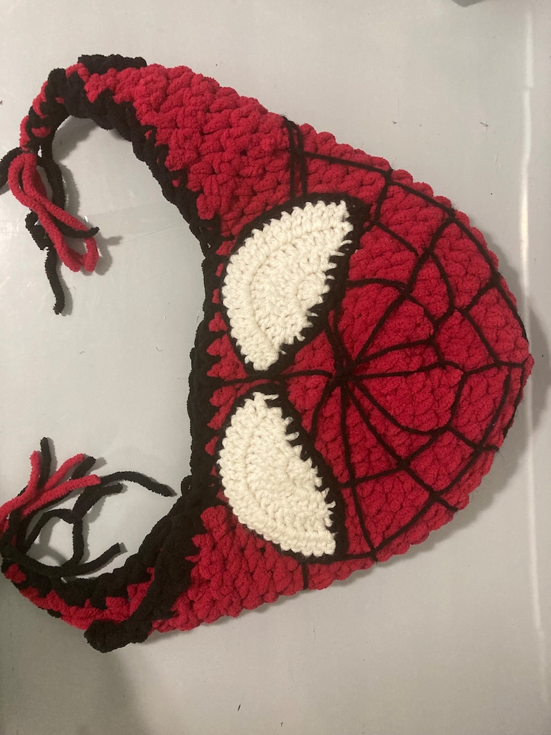 May include: A handmade crochet hat in the style of Spider-Man's mask. The hat features a red base with a black web design and white eye details. Black yarn is used for the edges and ties.