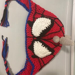 May include: A handmade crochet hat in the style of Spider-Man. The hat is primarily red with black web detailing, blue trim, and white eye shapes. A braided tassel of blue, black, and red yarn hangs from the hat.