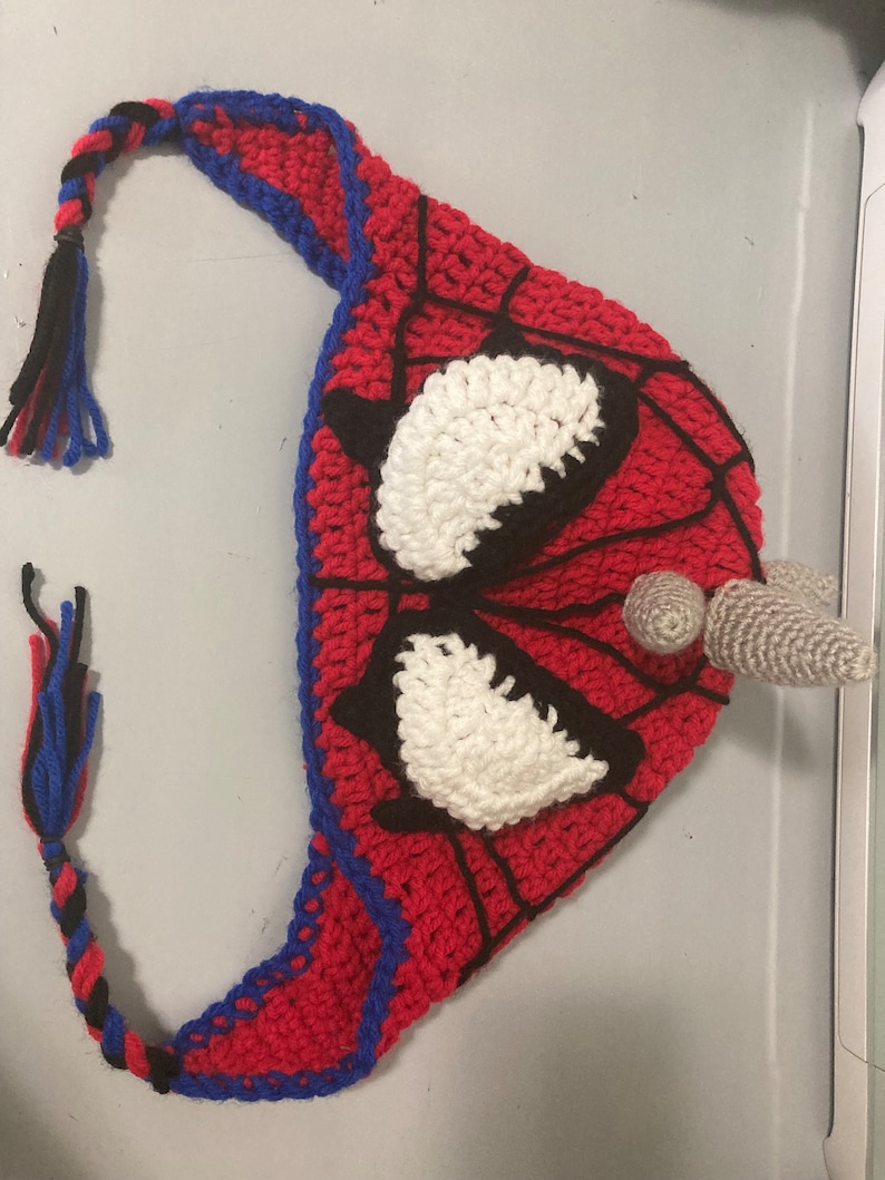 May include: A handmade crochet hat in the style of Spider-Man. The hat is primarily red with black web detailing, blue trim, and white eye shapes. A braided tassel of blue, black, and red yarn hangs from the hat.