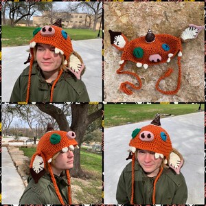 May include: A crocheted orange and brown monster hat with green and blue eyes, a pink nose, and white teeth. The hat has long orange and brown ties and floppy ears.