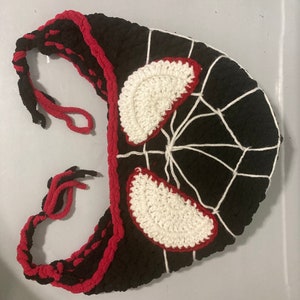 May include: A black and red crocheted mask with white accents. The mask features white crocheted eye shapes outlined in red, and a white web design. The edges are trimmed in red, and it has braided ties.
