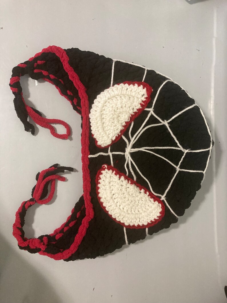 May include: A black and red crocheted mask with white accents. The mask features white crocheted eye shapes outlined in red, and a white web design. The edges are trimmed in red, and it has braided ties.