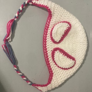 May include: A white crocheted hat with pink trim and accents. The hat features two pink-outlined, oval-shaped details and a braided strap with pink, white, and blue yarn. The hat is handmade and suitable for various occasions.