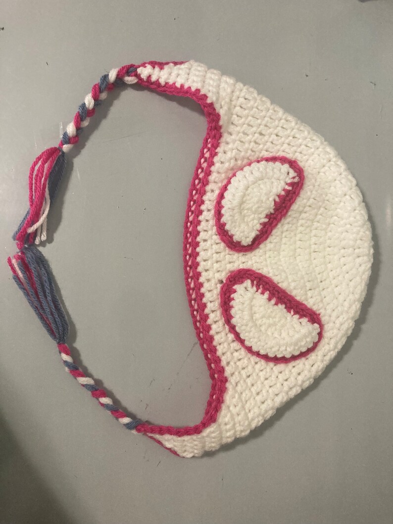 May include: A white crocheted hat with pink trim and accents. The hat features two pink-outlined, oval-shaped details and a braided strap with pink, white, and blue yarn. The hat is handmade and suitable for various occasions.