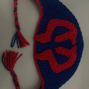 May include: A handmade crochet bag in blue with a red design. The bag has a braided strap with red and blue yarn and a tassel. The bag's design is a red, stylized letter. The bag is made of yarn.