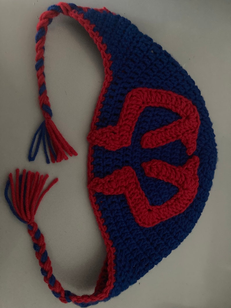 May include: A handmade crochet bag in blue with a red design. The bag has a braided strap with red and blue yarn and a tassel. The bag's design is a red, stylized letter. The bag is made of yarn.