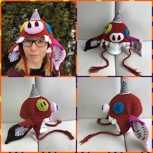 May include: A red, white, and black crocheted hat with a pig face and horns. The hat has a long tail with a tassel. The hat is on a white mannequin head.