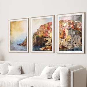 May include: Three framed watercolor paintings of a colorful Italian village on a cliff overlooking the ocean. The paintings are hung on a white wall above a white sofa with pillows.