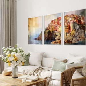 May include: Three framed watercolor paintings of a colorful Italian village on a cliff overlooking the ocean. The paintings are in white frames and are hung on a white wall.