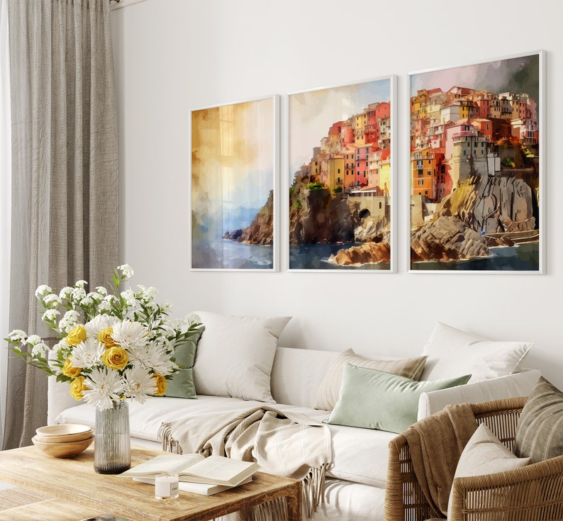 May include: Three framed watercolor paintings of a colorful Italian village on a cliff overlooking the ocean. The paintings are in white frames and are hung on a white wall.
